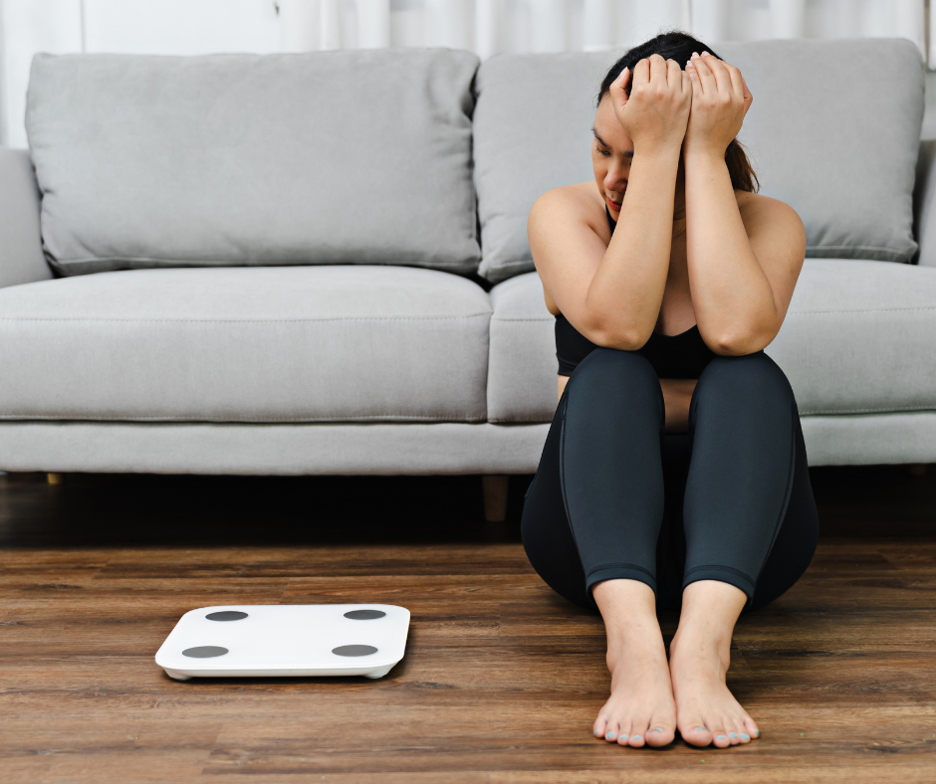 Woman sits on floor next to scale while thinking about the link between alcohol and weight gain