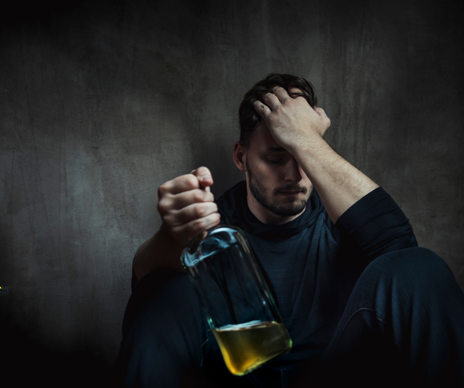 distraught young man leaning against wall in dimly lit room with head in his left hand and mostly empty liquor bottle in his right hand